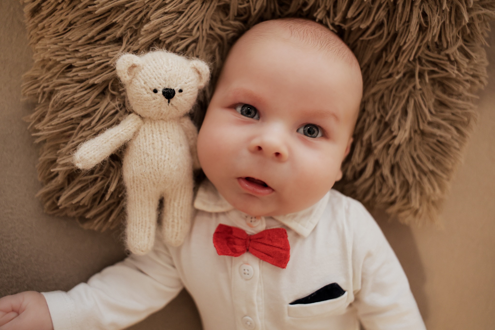 Niemowlę w białej koszuli z czerwoną muszką leży na puszystym tle obok pluszowego misia. Ujęcie z góry, miękkie światło.