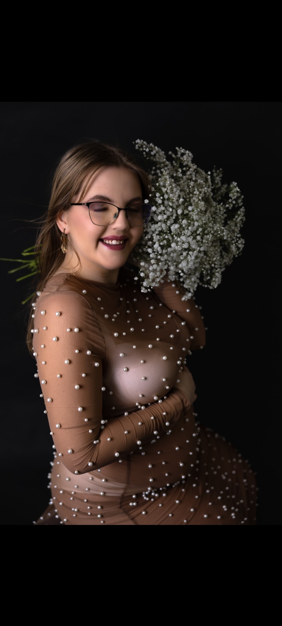 Uśmiechnięta kobieta w ciąży, w beżowej sukience z perełkami, pozuje z bukietem gipsówki na ciemnym tle. Okulary i kolczyki dodają elegancji.