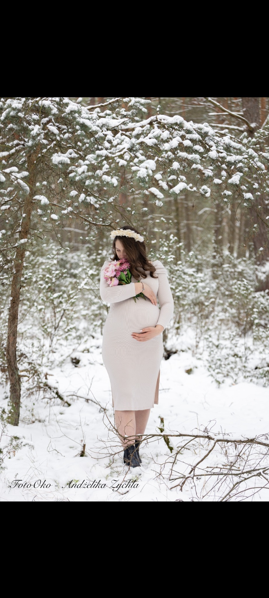Kobieta w ciąży w beżowej sukience, z wiankiem na głowie i bukietem różowych kwiatów, pozuje w zimowym lesie. Delikatne światło podkreśla naturalność sesji.