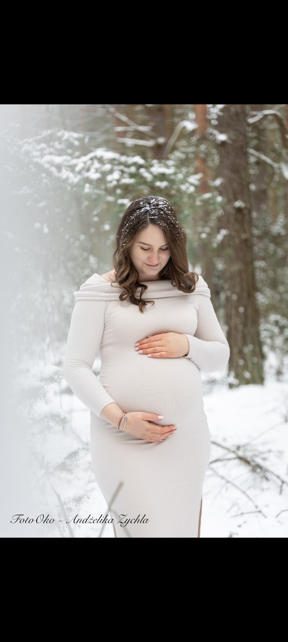 Kobieta w dopasowanej sukience, z rękami na brzuchu, pozuje w zimowej scenerii. Na włosach widoczne płatki śniegu. Ujęcie z delikatnym rozmyciem tła.
