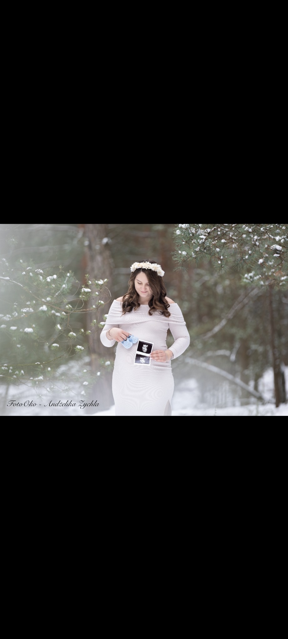 Kobieca sesja ciążowa w zimowej scenerii Kraszkowic. Kobieta w białej sukience trzyma zdjęcia USG i niebieskie buciki. Włosy zdobi wianek z białych kwiatów.