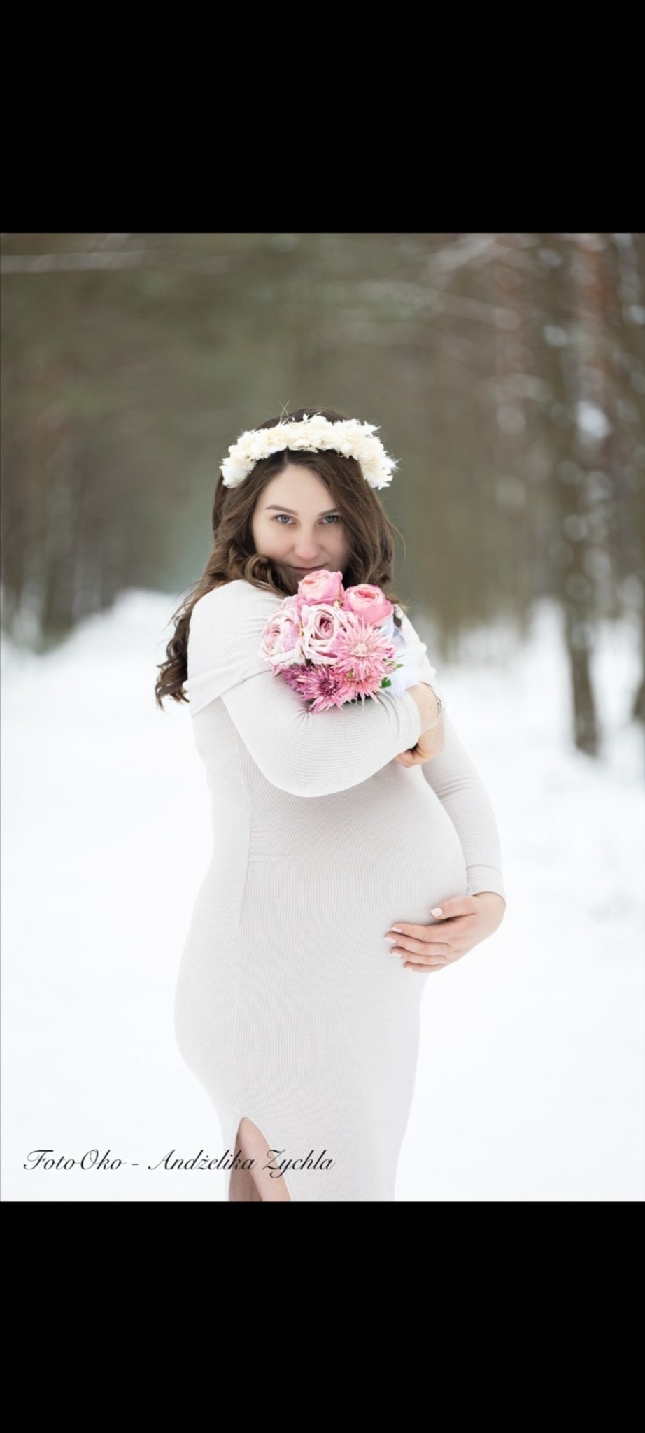 Kobieta w ciąży w białej sukience i wianku na głowie, trzymająca bukiet różowych kwiatów na tle zimowego krajobrazu. Ujęcie z delikatnym uśmiechem i skupieniem na brzuszku.
