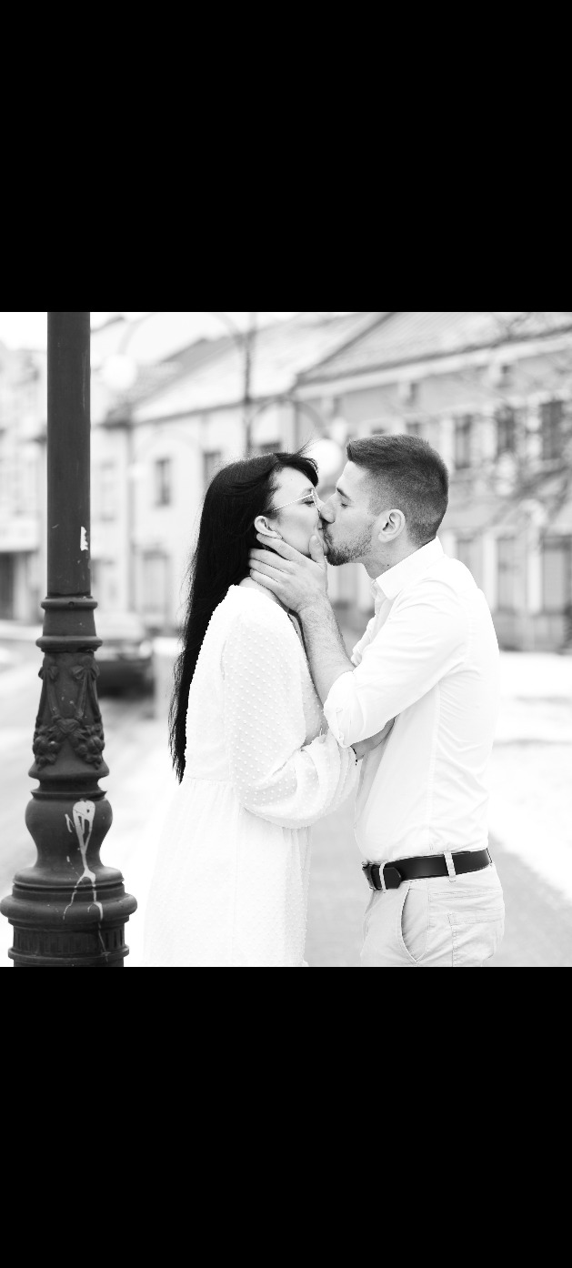 Czarno-białe zdjęcie całującej się pary na tle miejskiej uliczki z latarnią. Kobieta w białej sukience, mężczyzna w koszuli. Romantyczny kadr z sesji narzeczeńskiej.