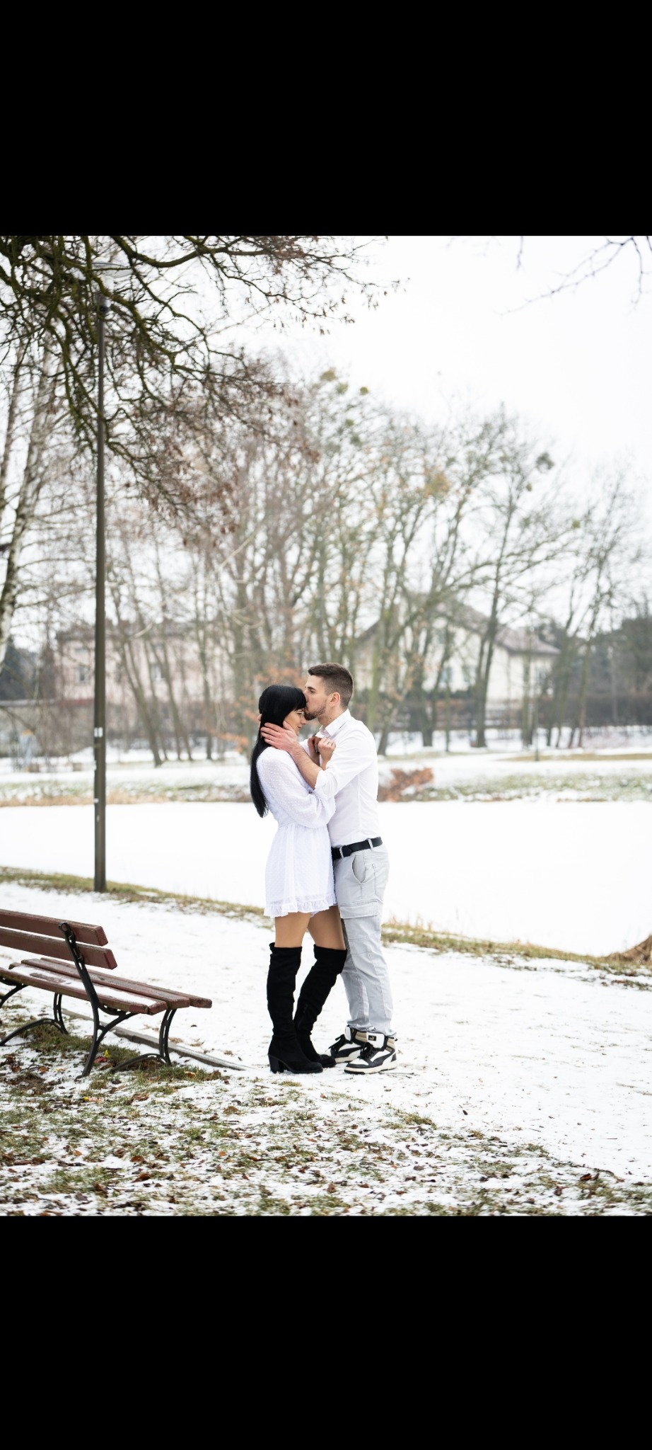 Zimowa sesja narzeczeńska w parku: mężczyzna całuje kobietę w czoło, oboje ubrani na biało, na tle ośnieżonego krajobrazu i ławek. Ujęcie pełne czułości i naturalności.