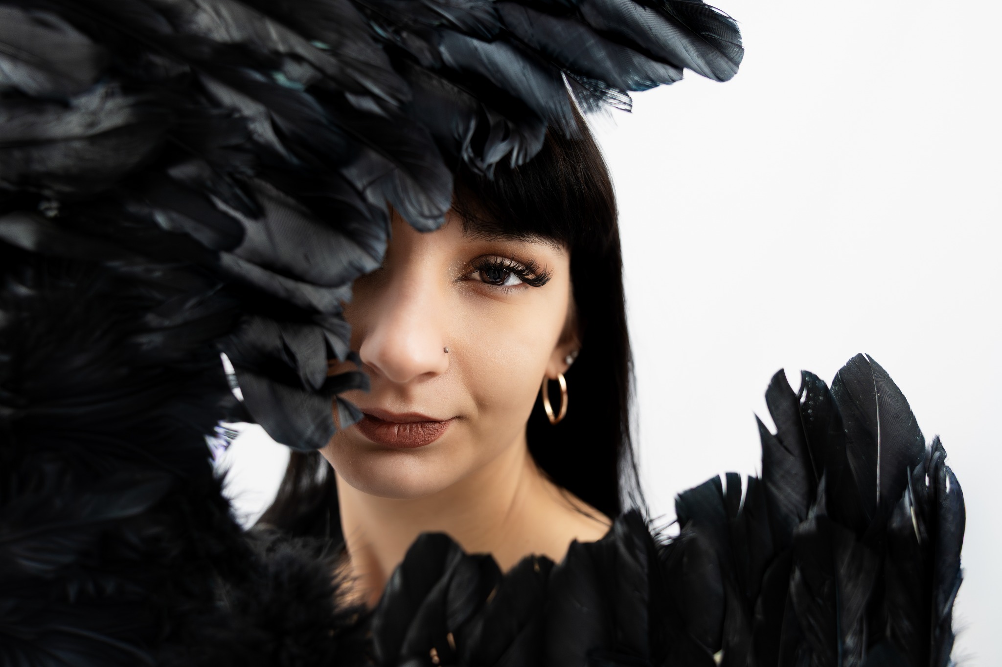 Portret kobiety z czarnymi piórami, złote kolczyki koła, piercing nosa, wyraziste rzęsy, ciemne włosy z grzywką na białym tle. Artystyczna sesja.