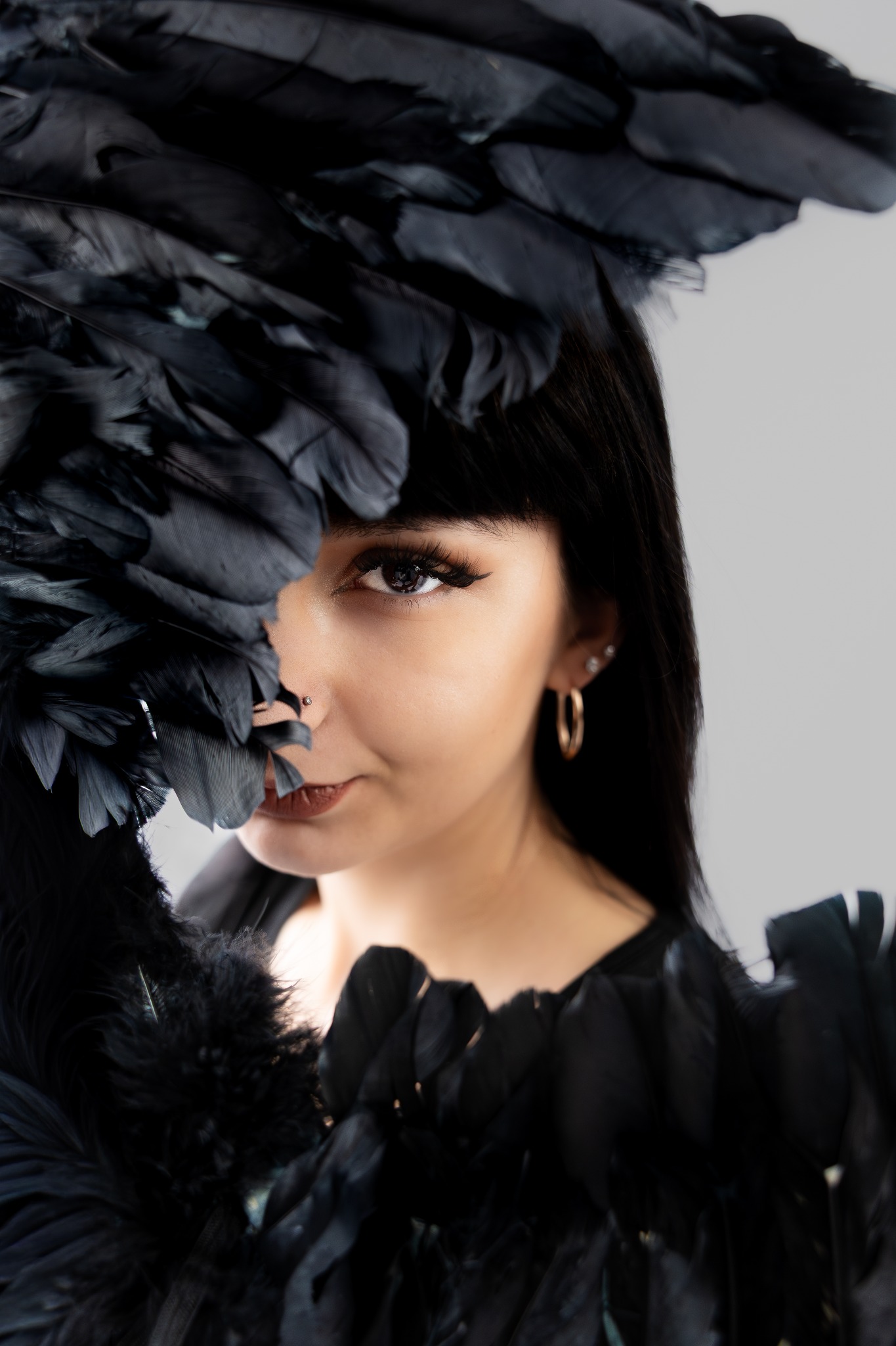 Portret kobiety z czarnymi piórami zasłaniającymi część twarzy, z widocznym piercingiem nosa i złotymi kolczykami na tle jasnoszarego studia.