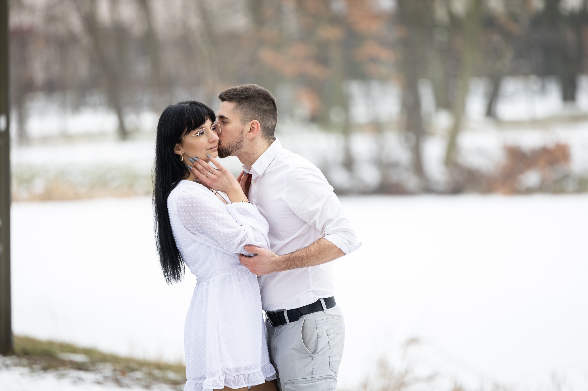 Zimowa sesja narzeczeńska: mężczyzna całuje kobietę w okularach, oboje ubrani na biało, na tle ośnieżonego parku z rozmytymi drzewami.