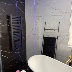 Kacper Zbroja - Elegancka łazienka z czarno-białymi płytkami imitującymi marmur, podświetleniem LED, wolnostojącą wanną i grzejnikiem drabinkowym. Nowoczesny design z akcentami złota.