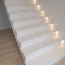 GlamourStone - Białe schody z subtelnym oświetleniem LED pod stopniami, widok z dołu, minimalistyczny design, jasne drewniane panele podłogowe, nowoczesne wykończenie.