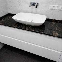 GlamourStone - Łazienka z czarnym blatem kamiennym o marmurkowym wzorze, białą umywalką nablatową i szafką z dwiema szufladami. Minimalistyczny design, płytki imitujące marmur na ścianie.
