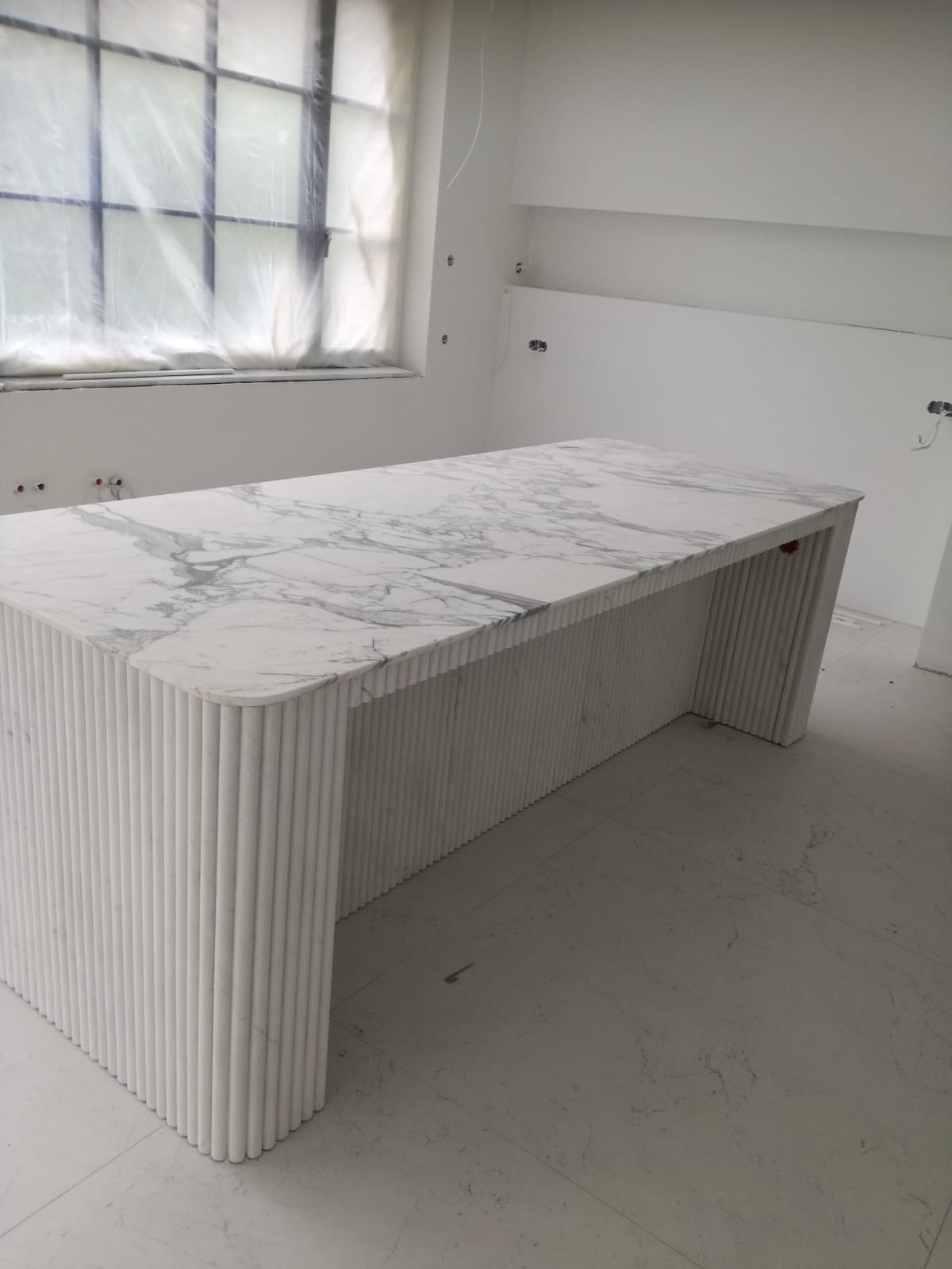 Biały blat z marmuru Carrara, wsparty na ryflowanych, cylindrycznych nogach. Minimalistyczny design, surowe wnętrze w trakcie wykończenia. Widok z perspektywy podłogi.