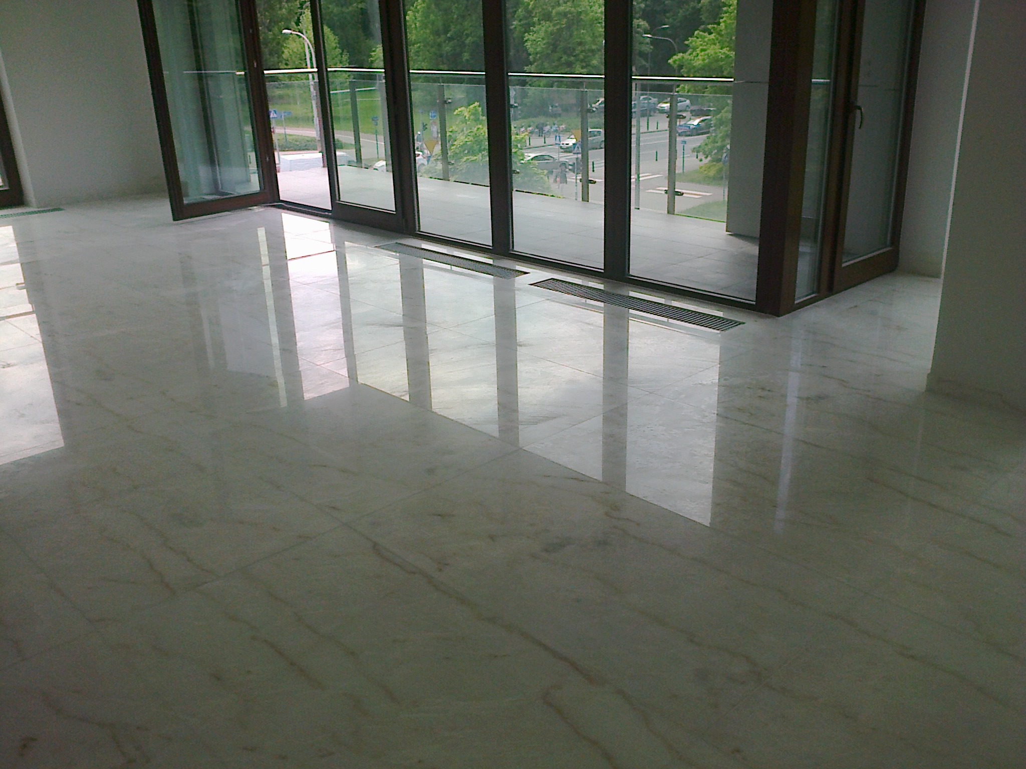 Błyszcząca, kremowa posadzka z kamiennych płyt w nowoczesnym wnętrzu z dużymi, przesuwnymi drzwiami balkonowymi i widokiem na zieleń miejską.