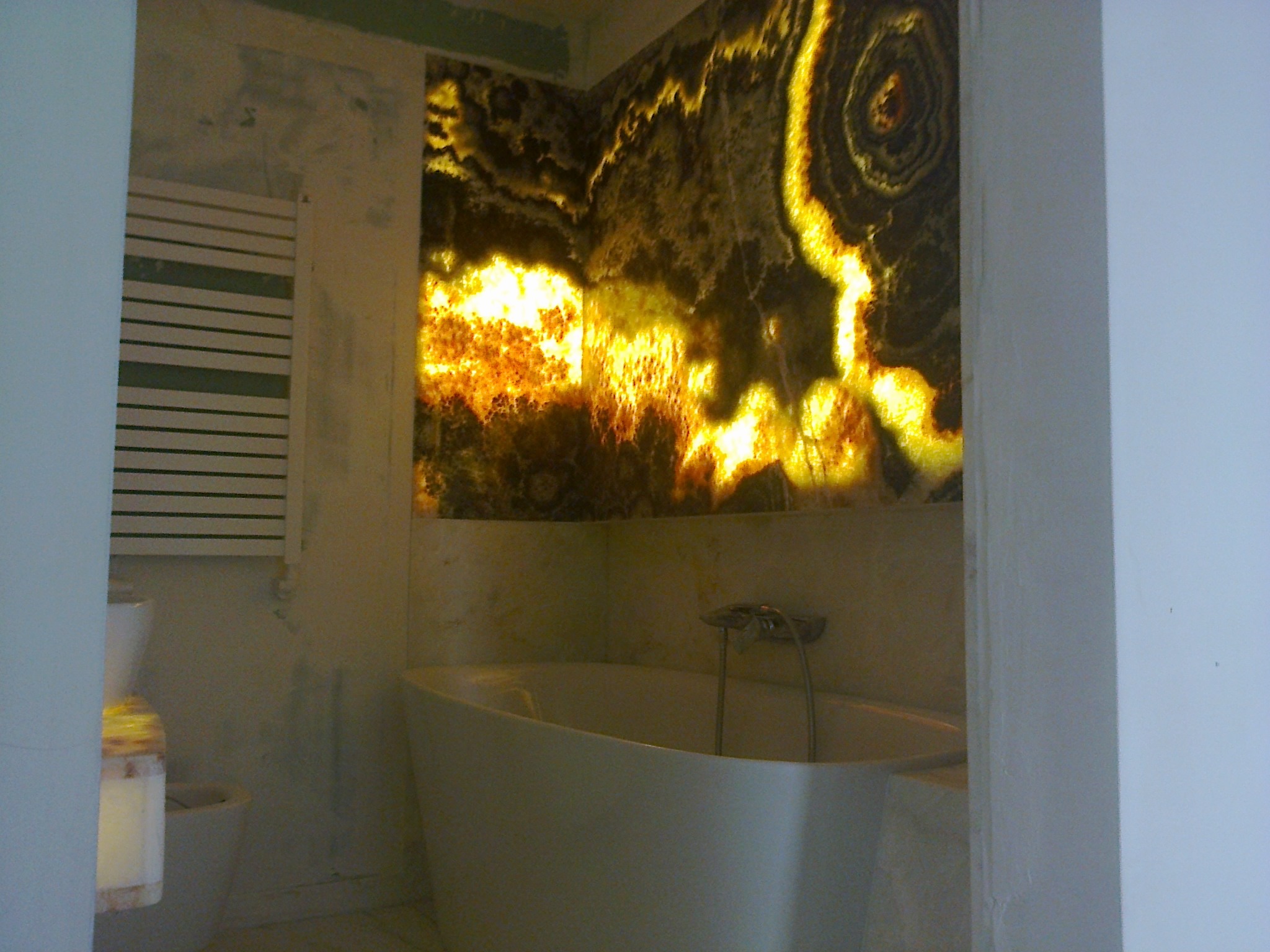 Podświetlona ściana z onyksu w łazience, z widoczną wanną i grzejnikiem. Unikalny wzór kamienia tworzy ciepłą, dekoracyjną atmosferę.