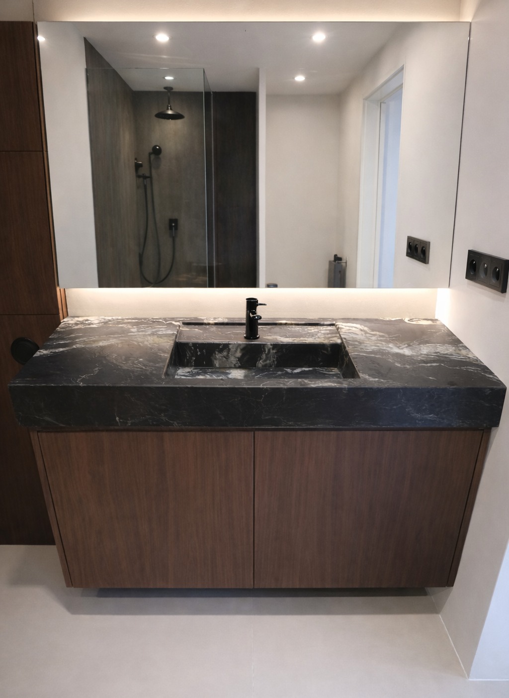 Elegancka łazienka z blatem z czarnego kamienia o marmurkowej fakturze, wpuszczona umywalka i czarna bateria. W tle lustro z oświetleniem LED i widok na kabinę prysznicową.