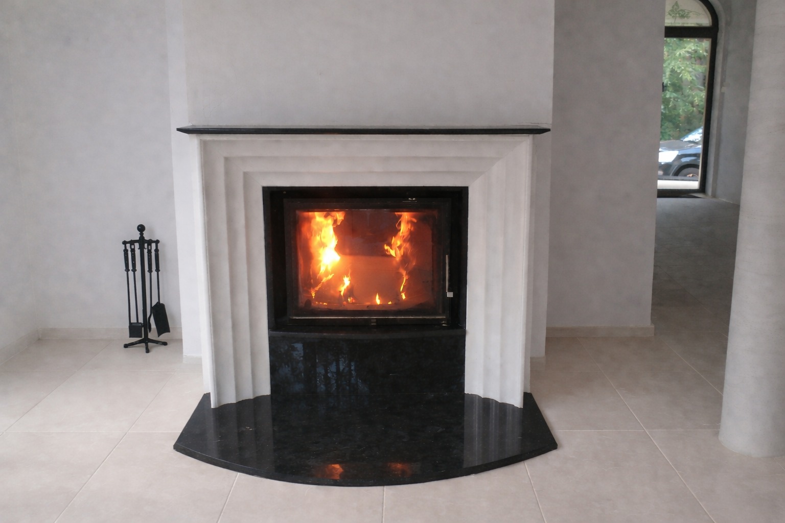 Nowoczesny kominek z czarnym kamiennym blatem i białym obramowaniem, płomienie wewnątrz paleniska, akcesoria kominkowe obok. Jasne płytki podłogowe.