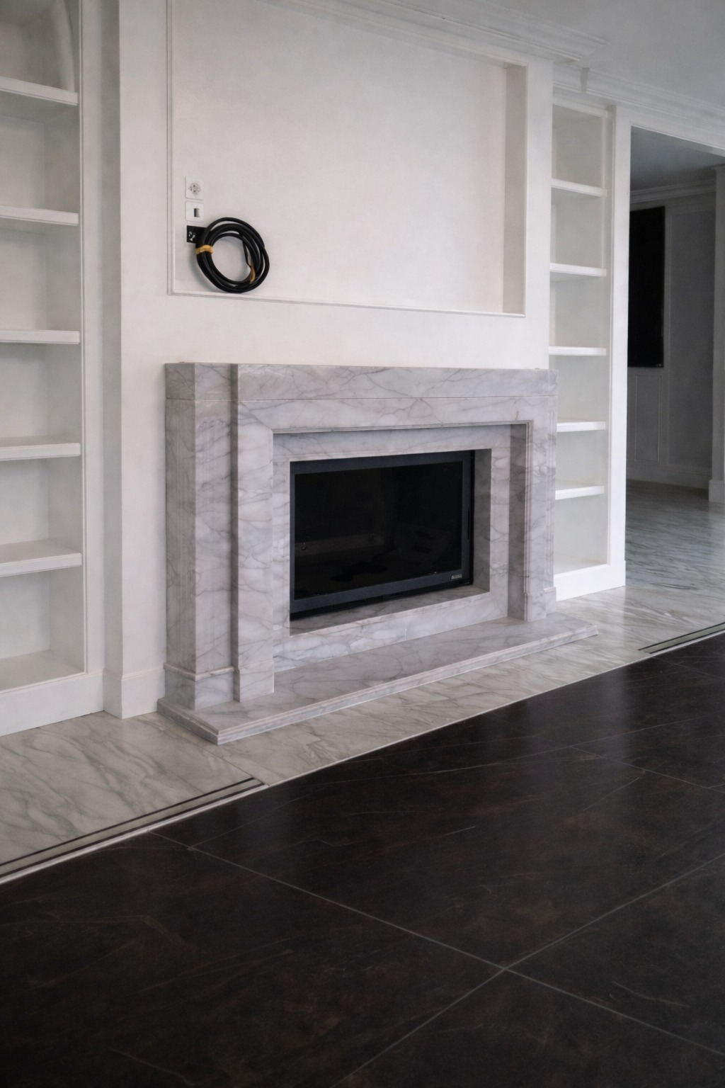 Elegancki marmurowy kominek z czarnym wkładem, wkomponowany w białą zabudowę z półkami. Podłoga wyłożona płytkami imitującymi marmur i ciemny kamień.