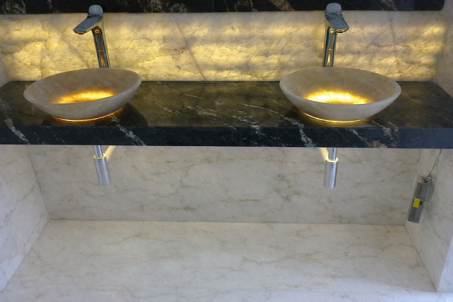 Dwie owalne umywalki z podświetleniem LED na czarnym, marmurkowym blacie. Tło stanowi podświetlona ściana z jasnego kamienia z wyraźnym użyleniem.