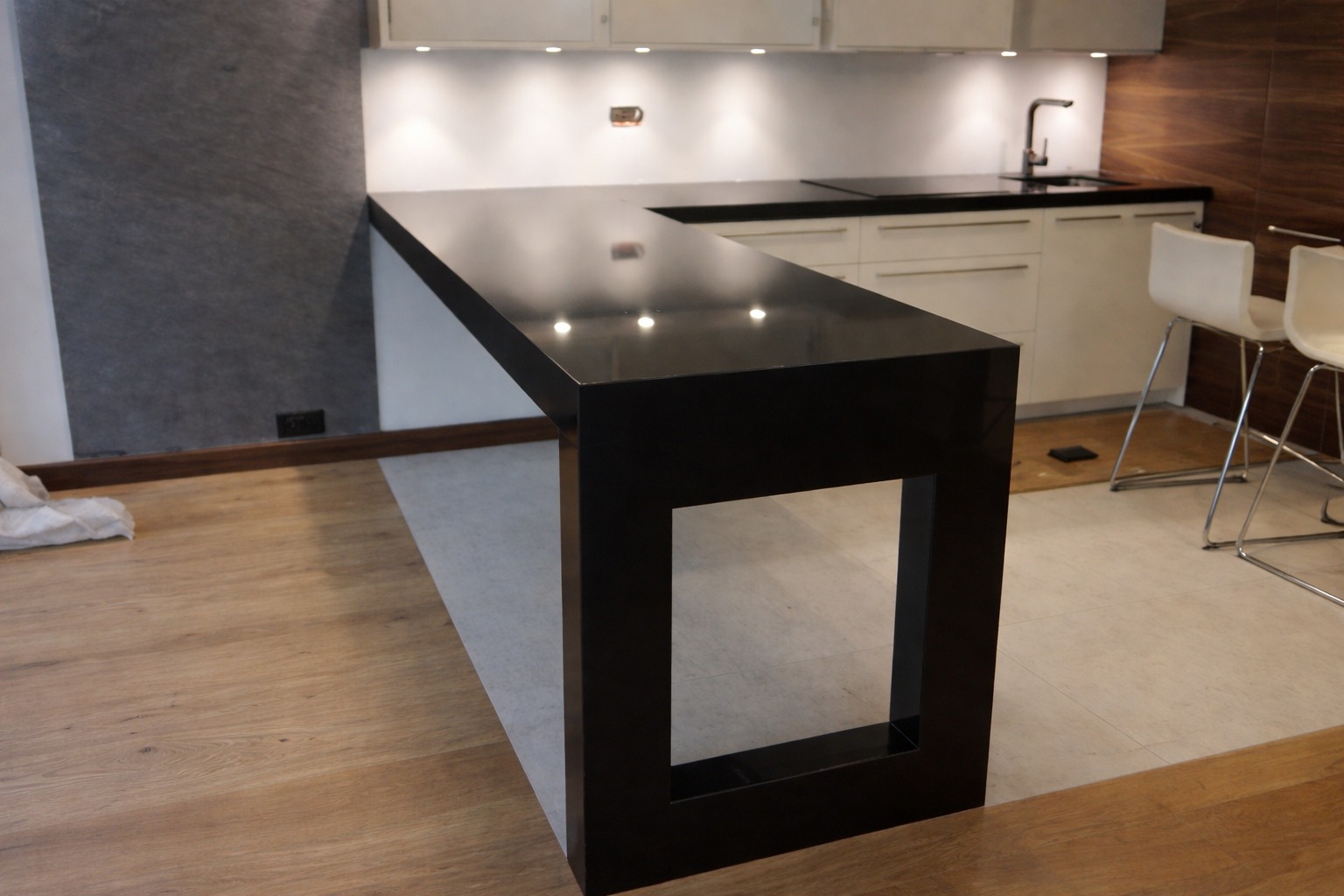 Czarny, lśniący blat kamienny w nowoczesnej kuchni z białymi szafkami i drewnianą podłogą. Widoczne dwa białe krzesła barowe i fragment ściany z szarą okładziną.