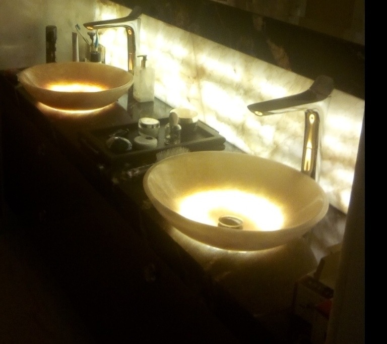 Dwie owalne umywalki z kamienia, podświetlone od spodu, z nowoczesną armaturą na blacie z kamienia. W tle ściana z podświetlanego kamienia.