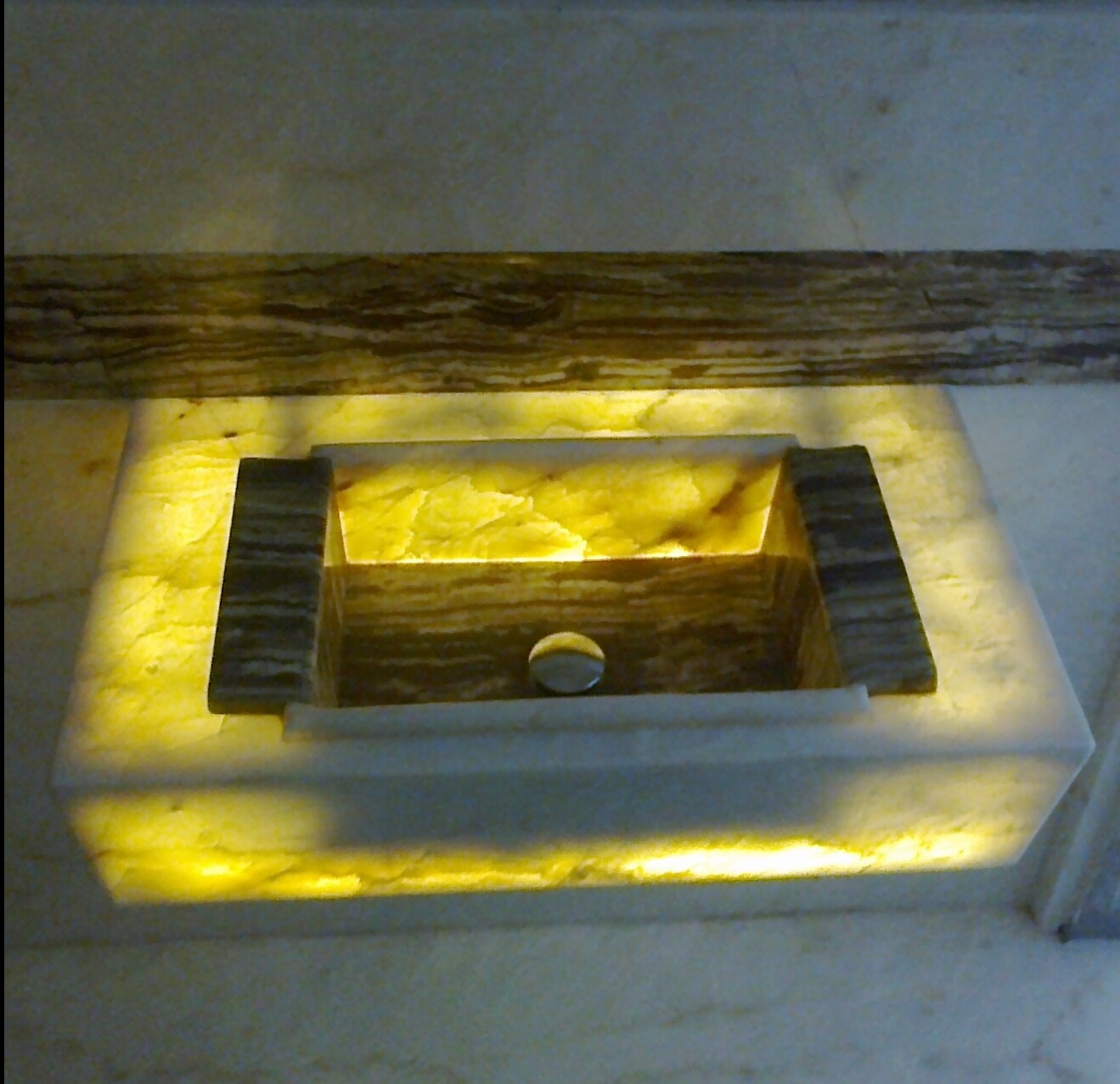 Podświetlana, prostokątna umywalka z onyksu z ciemnymi elementami wykończeniowymi, widoczna od góry, z bliska. Ciepłe światło podkreśla unikalny wzór kamienia.