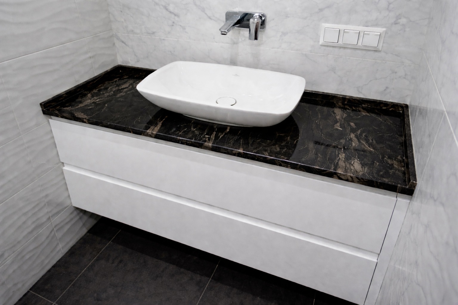 Łazienka z czarnym blatem kamiennym o marmurkowym wzorze, białą umywalką nablatową i szafką z dwiema szufladami. Minimalistyczny design, płytki imitujące marmur na ścianie.