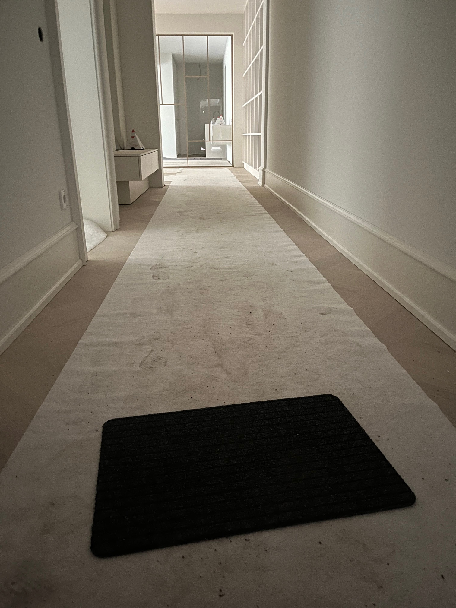 Jasny korytarz w trakcie remontu, podłoga zabezpieczona folią ochronną, widoczne listwy przypodłogowe i drzwi z przeszkleniami w głębi.