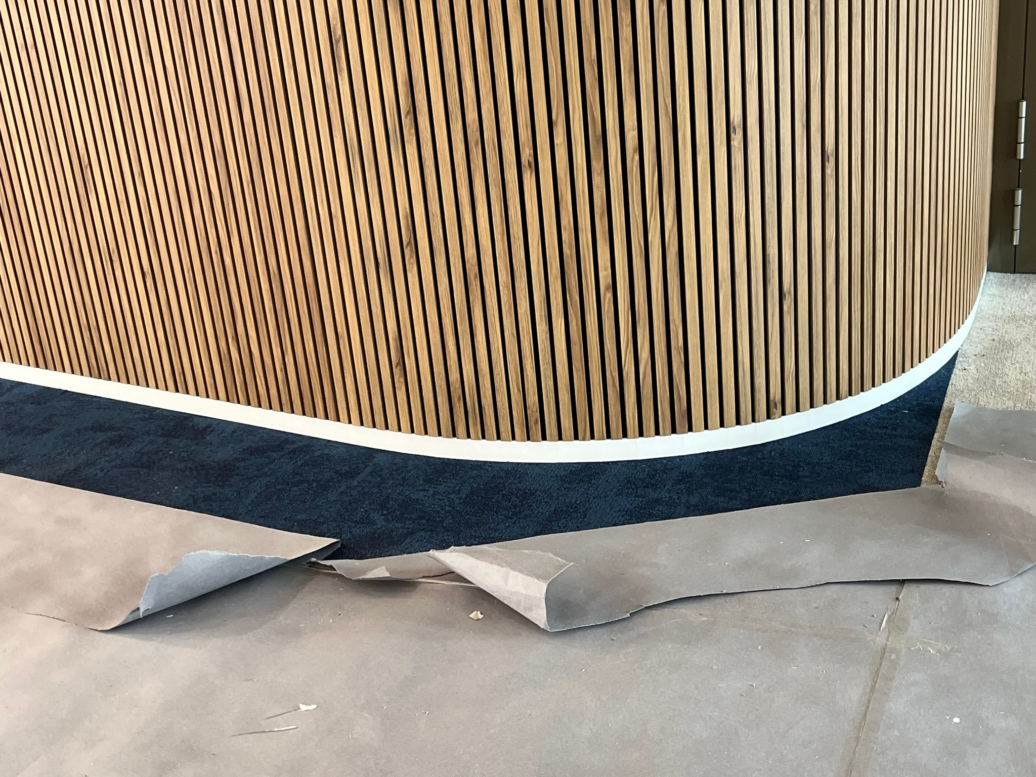 Zaokrąglona ściana z drewnianych paneli lamelowych w trakcie montażu, widoczne zabezpieczenia podłogi i fragment wykładziny. Detal wykończenia wnętrza.