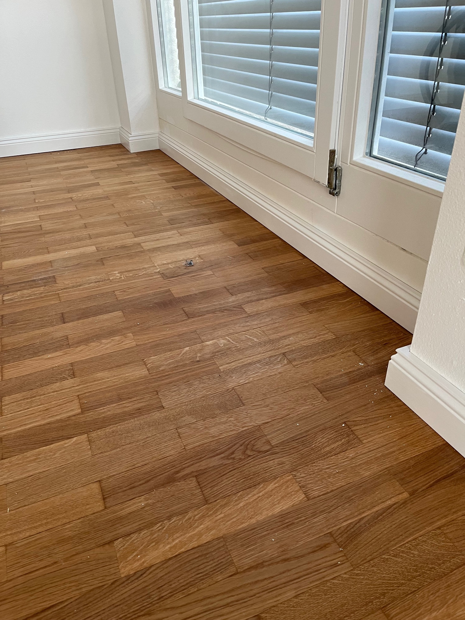 Ukończona podłoga z paneli drewnianych w jasnym wnętrzu z białymi listwami przypodłogowymi i oknem z żaluzjami. Widoczne drobne zabrudzenia po montażu.