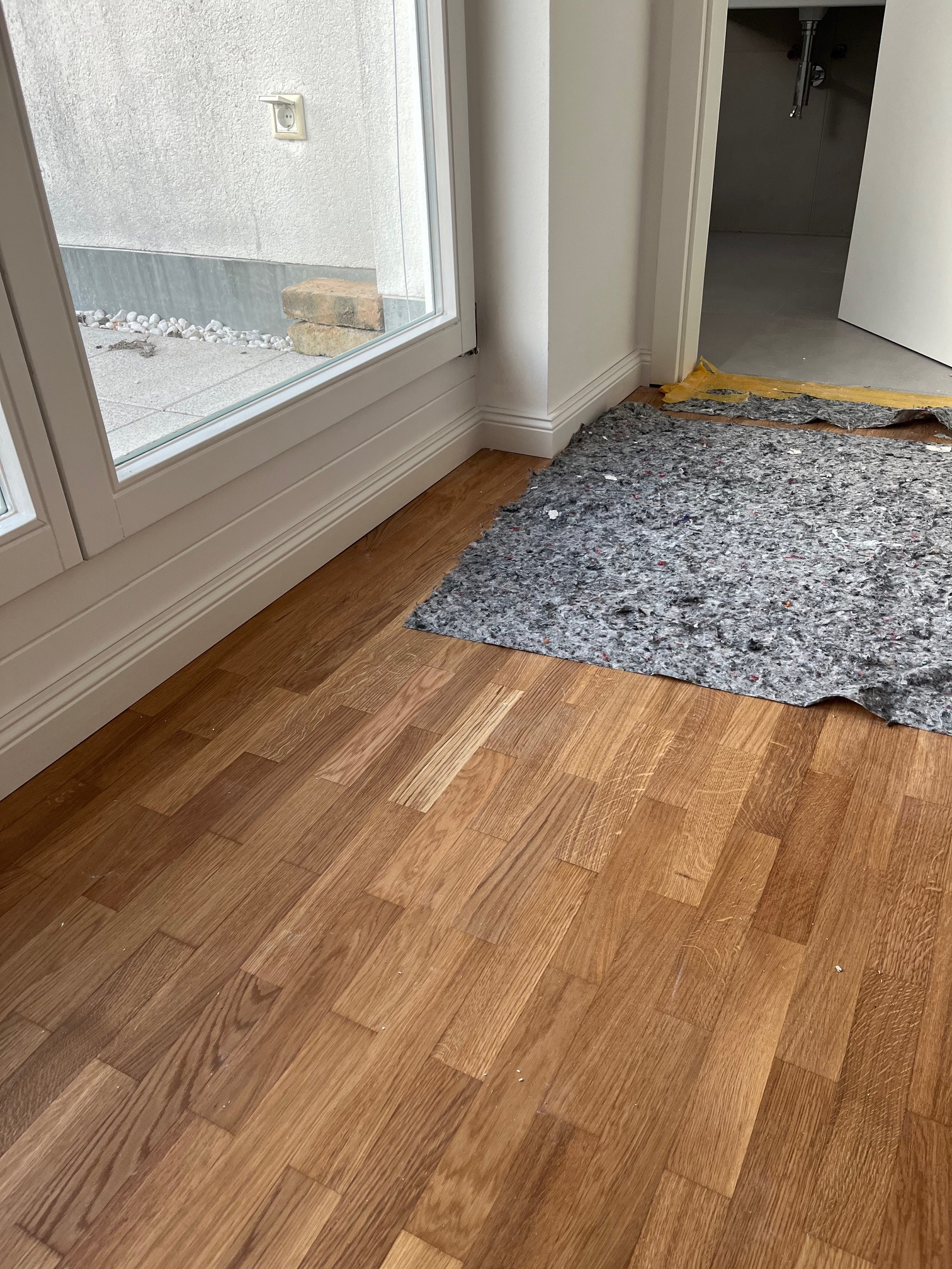 Fragment wnętrza z ułożoną drewnianą podłogą panelową, widoczna mata wygłuszająca przy progu i okno z widokiem na zewnątrz. Jasne ściany i listwy przypodłogowe.