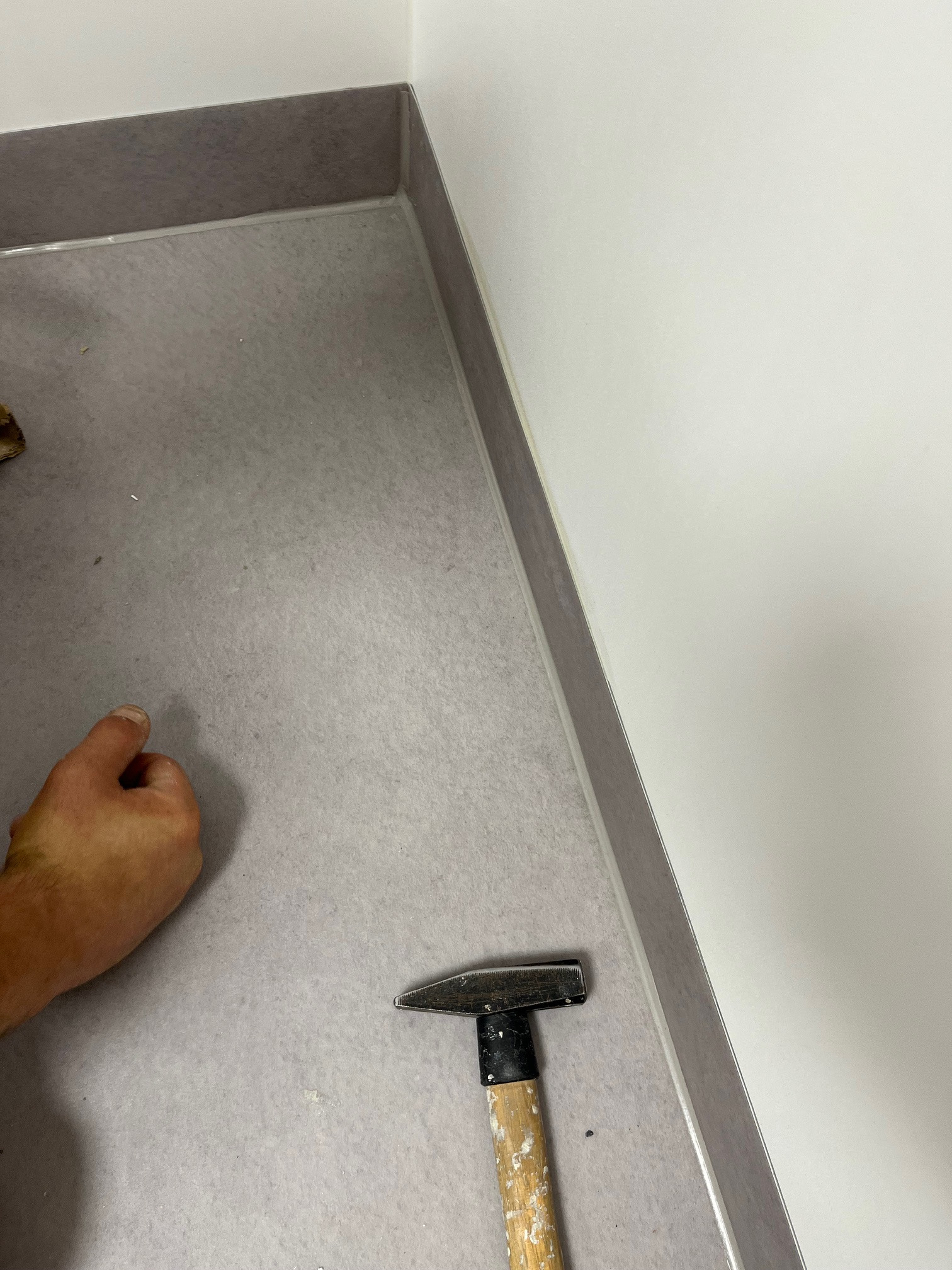 Szara podłoga z listwami przypodłogowymi, fragment dłoni i młotek. Widok z góry na etap montażu, detale wykończenia wnętrza.