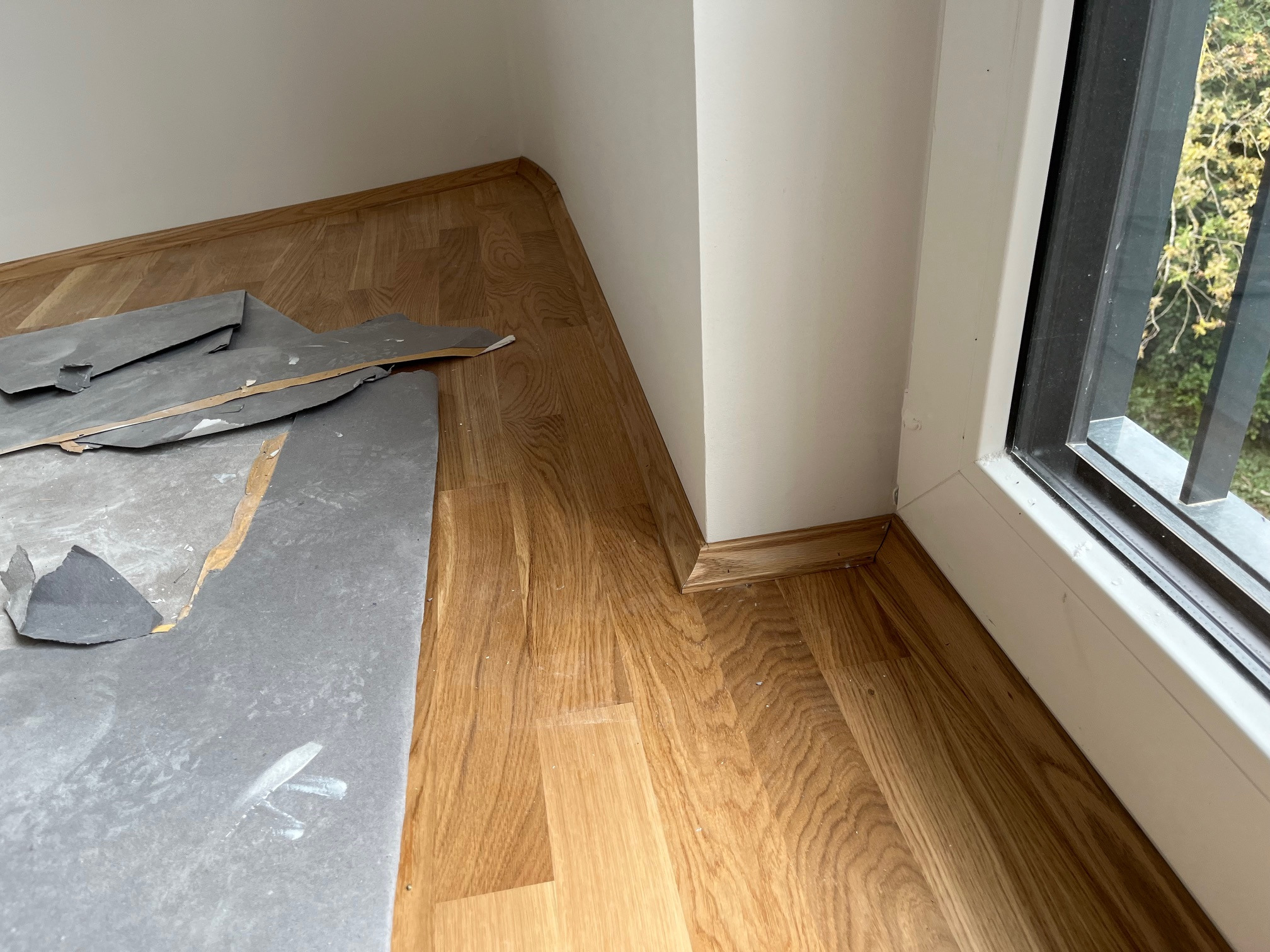 Fragment pokoju z drewnianą podłogą w trakcie remontu, widoczne panele i listwy przypodłogowe przy oknie. Częściowo ułożona podłoga, resztki materiałów obok.