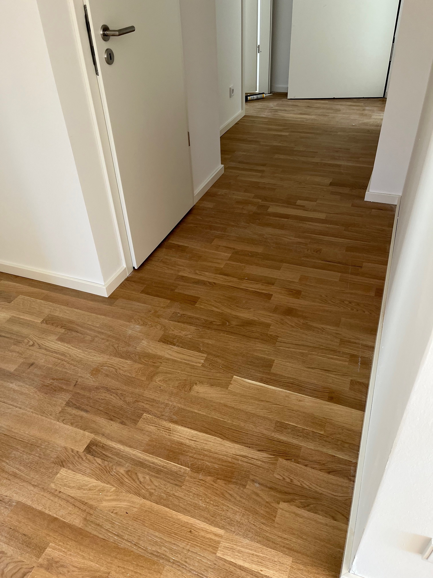 Jasne panele podłogowe ułożone w korytarzu z białymi ścianami i drzwiami. Widoczny fragment listwy przypodłogowej. Perspektywa z głębi pomieszczenia.