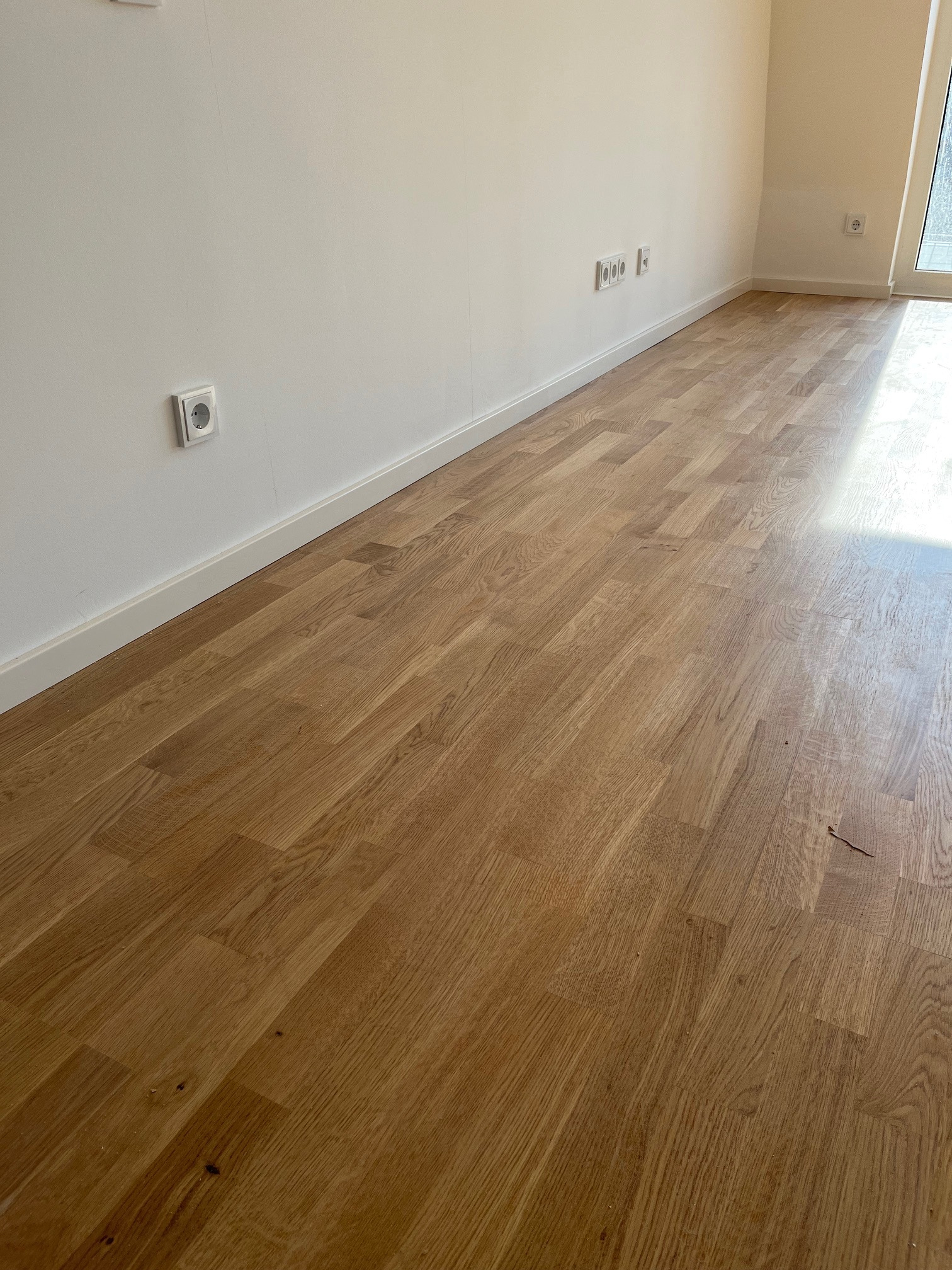 Jasne panele podłogowe ułożone w nowym, minimalistycznym wnętrzu. Widoczne listwy przypodłogowe i gniazdka elektryczne na ścianie. Naturalne światło podkreśla strukturę drewna.