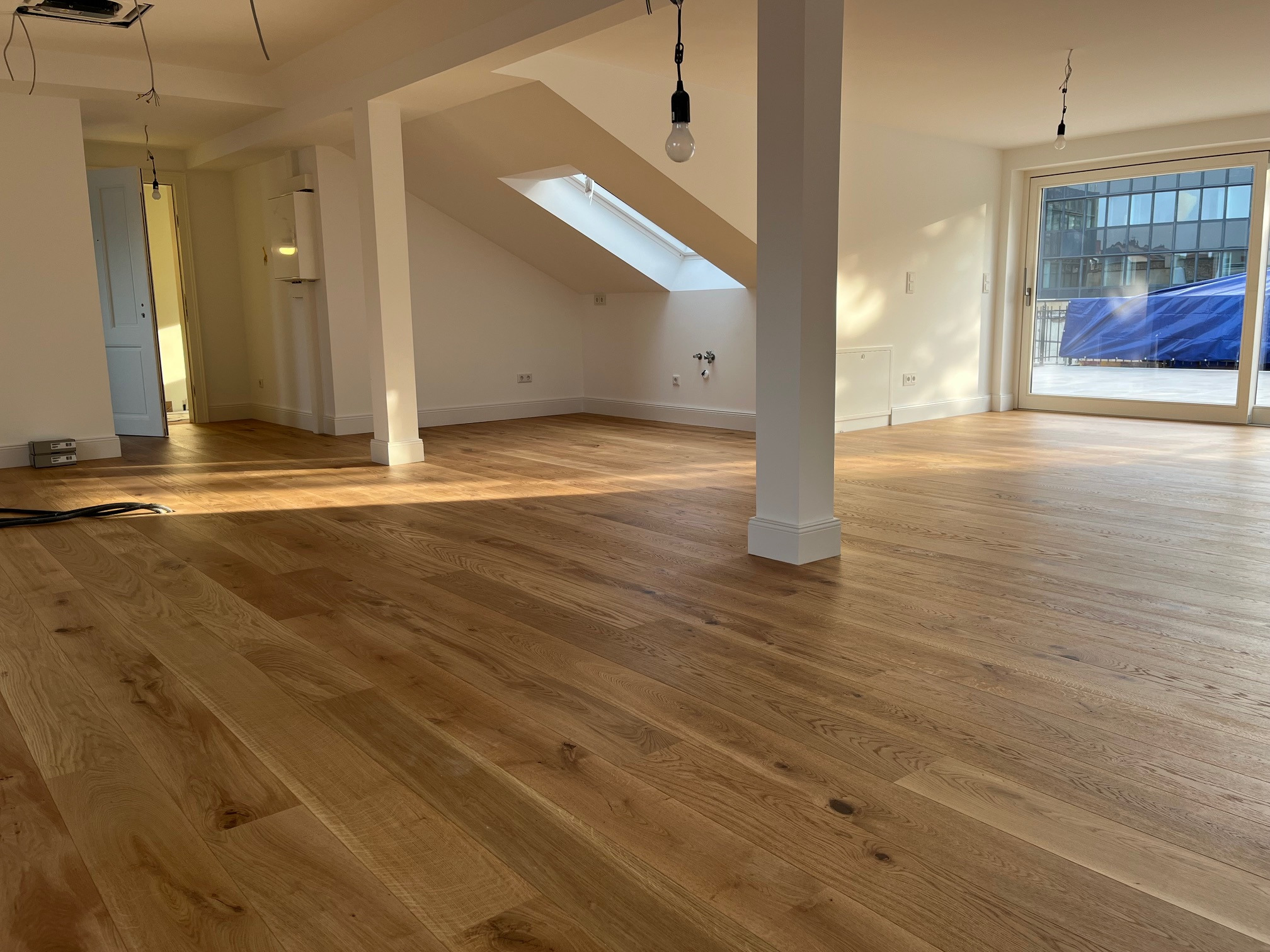 Jasne, drewniane panele podłogowe w przestronnym, odnowionym wnętrzu z białymi ścianami, kolumnami i oknem dachowym. Widoczne niedokończone instalacje.