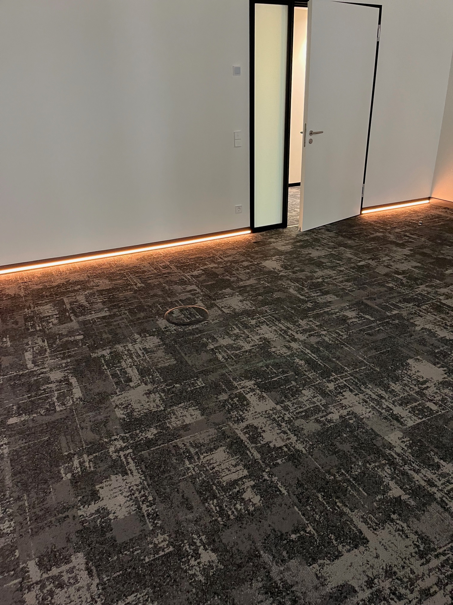 Nowoczesne wnętrze z szarą wykładziną dywanową w abstrakcyjny wzór, oświetleniem LED przy podłodze i otwartymi białymi drzwiami. Widoczny fragment podłogi z okrągłym otworem.