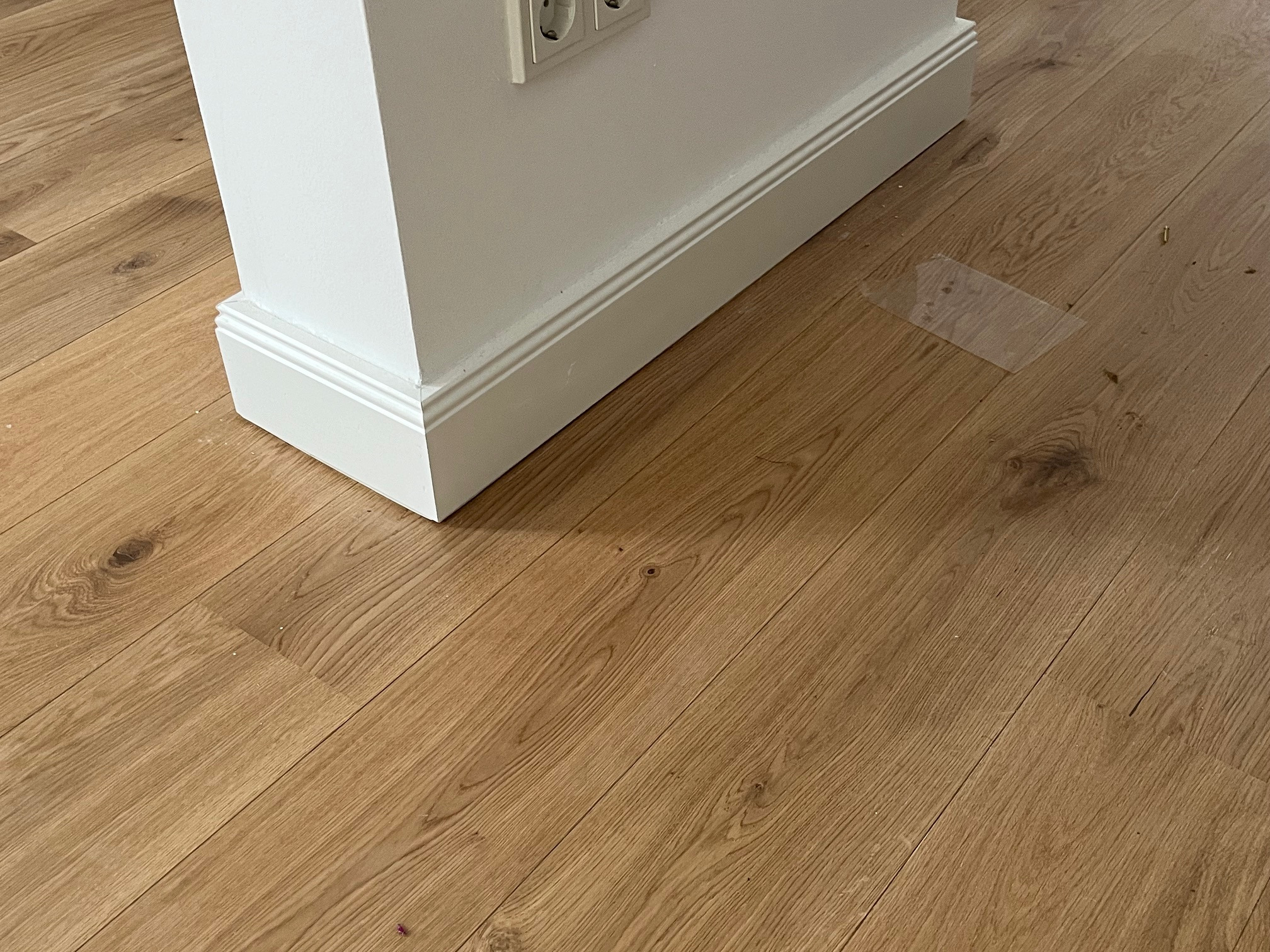 Jasne panele podłogowe z widoczną strukturą drewna, przy białej ścianie z listwą przypodłogową. Fragment gniazdka elektrycznego w górnym kadrze.