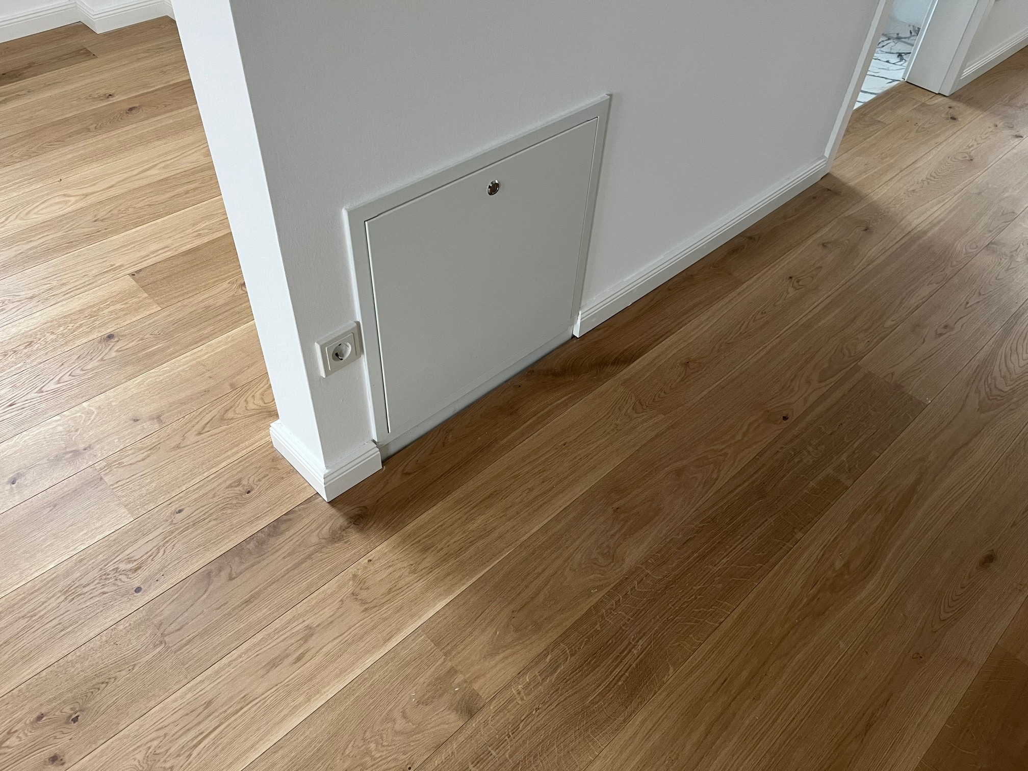 Jasne, drewniane panele podłogowe ułożone wzdłuż ściany z białą listwą przypodłogową i ukrytym włazem rewizyjnym. Widoczny fragment gniazdka elektrycznego.