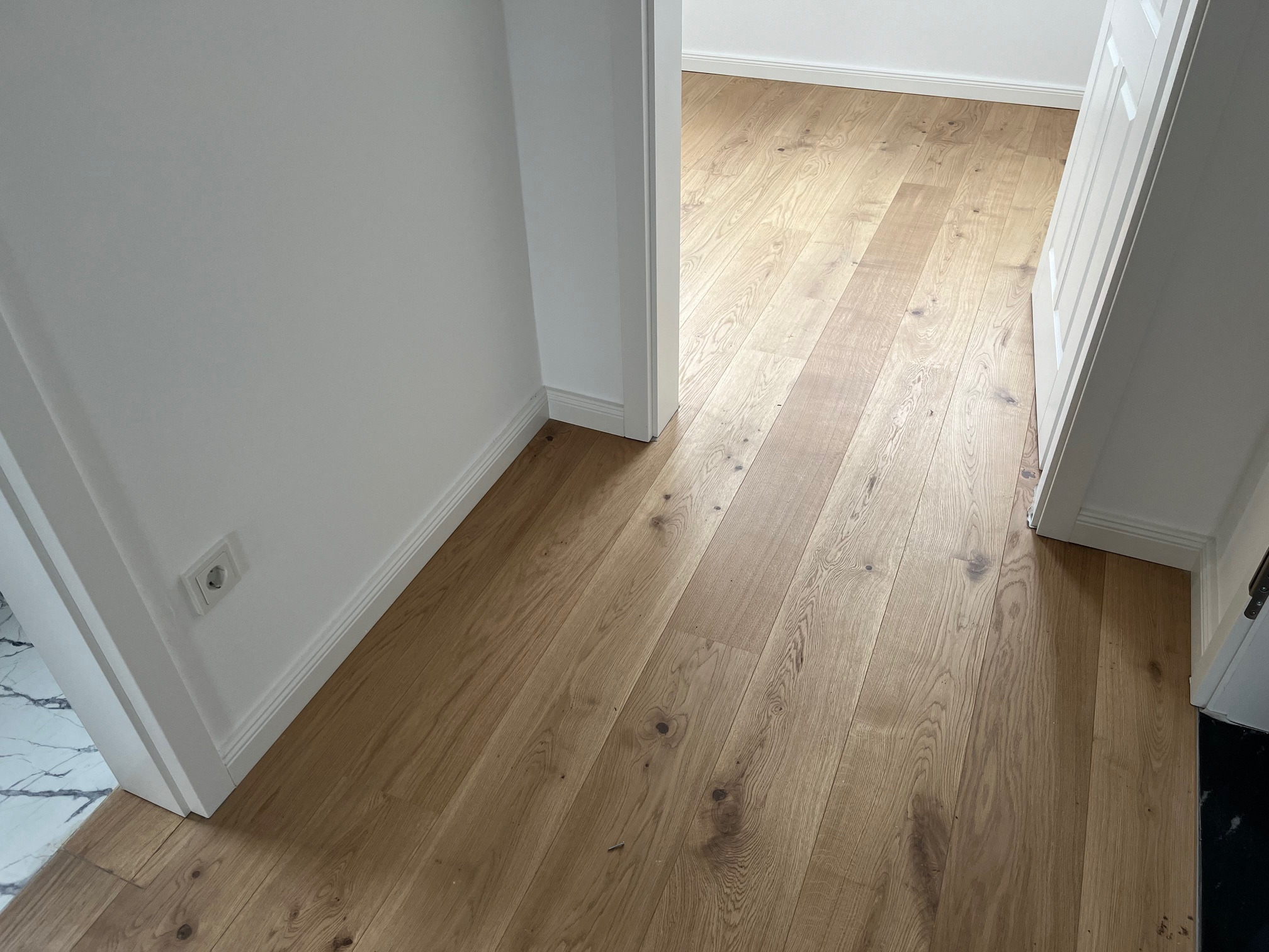 Jasne, drewniane panele podłogowe ułożone w korytarzu z białymi ścianami i listwami przypodłogowymi. Widoczny fragment marmurowej podłogi w przejściu.