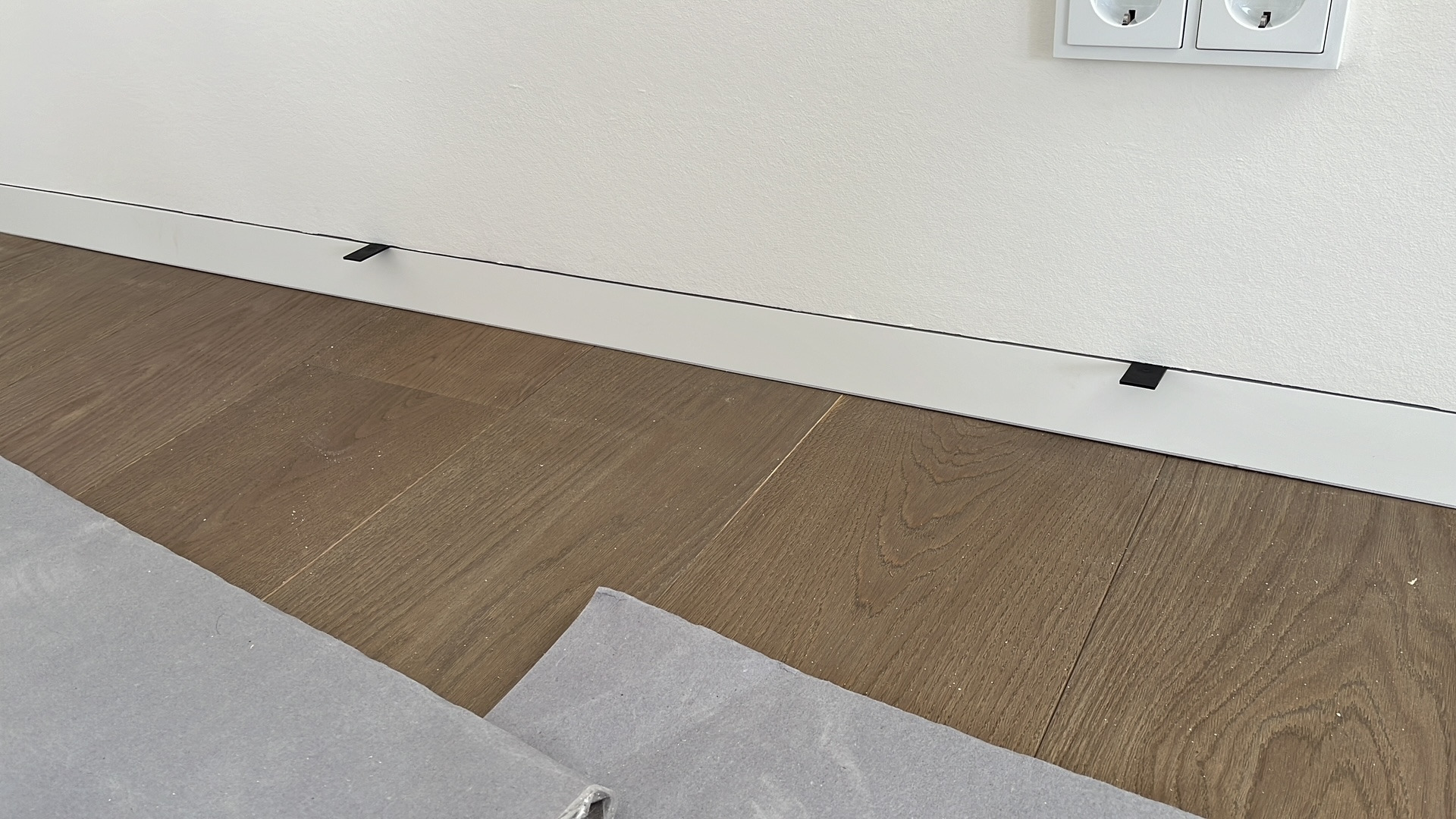 Biała listwa przypodłogowa z czarnymi klipsami montażowymi na brązowej podłodze z paneli. Fragment ściany z gniazdkiem elektrycznym w tle.