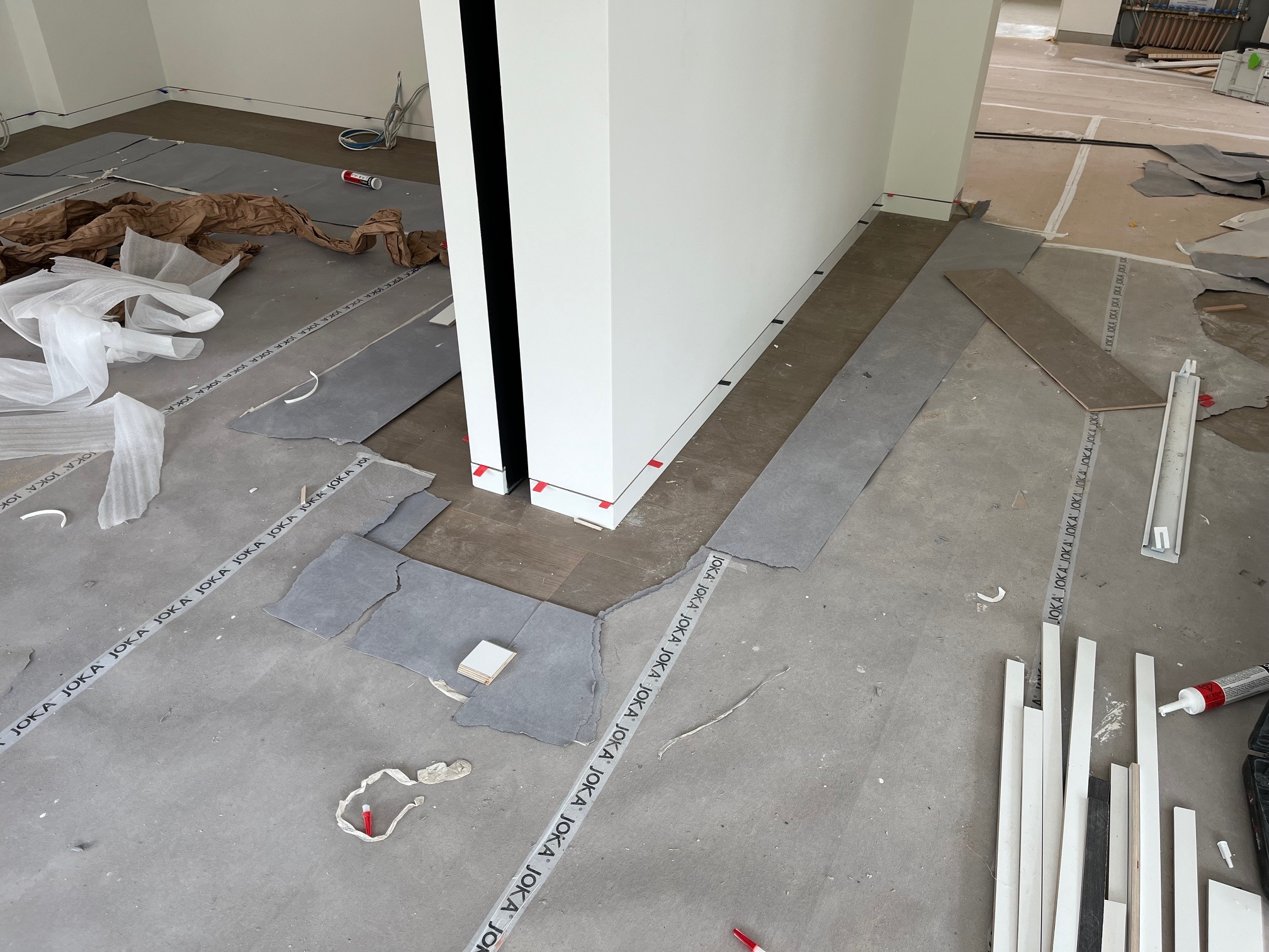 Fragment podłogi w trakcie układania paneli, widoczne podkłady, listwy przypodłogowe i narzędzia. Częściowo ułożone panele przy białej ścianie.