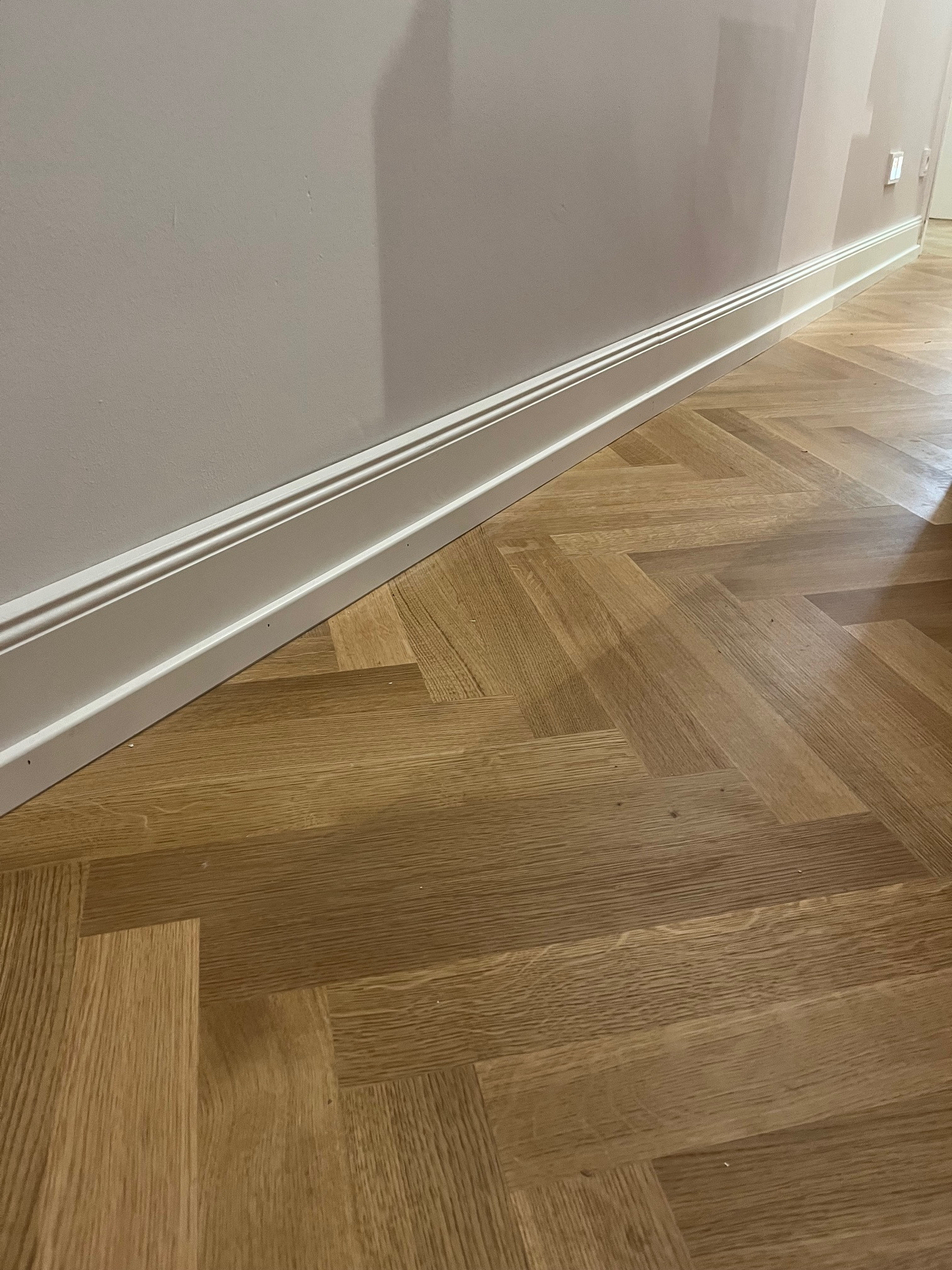 Parkiet ułożony w jodełkę, z widocznymi listwami przypodłogowymi w kolorze białym, zamontowanymi wzdłuż ściany pomalowanej na jasny odcień.