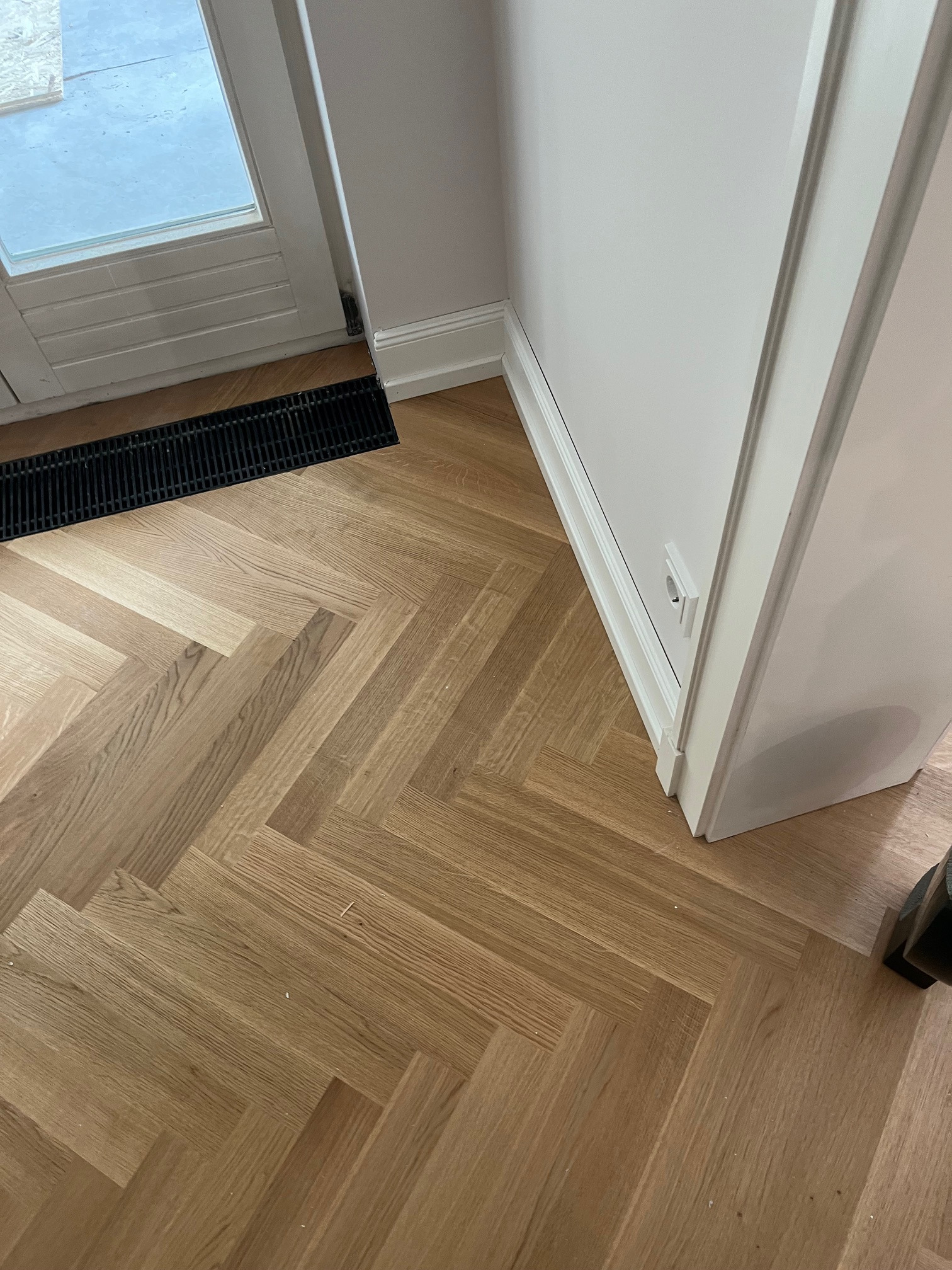 Parkiet ułożony w jodełkę francuską, widoczny fragment drzwi z kratką wentylacyjną i biała listwa przypodłogowa. Jasne drewno i minimalistyczne wykończenie.