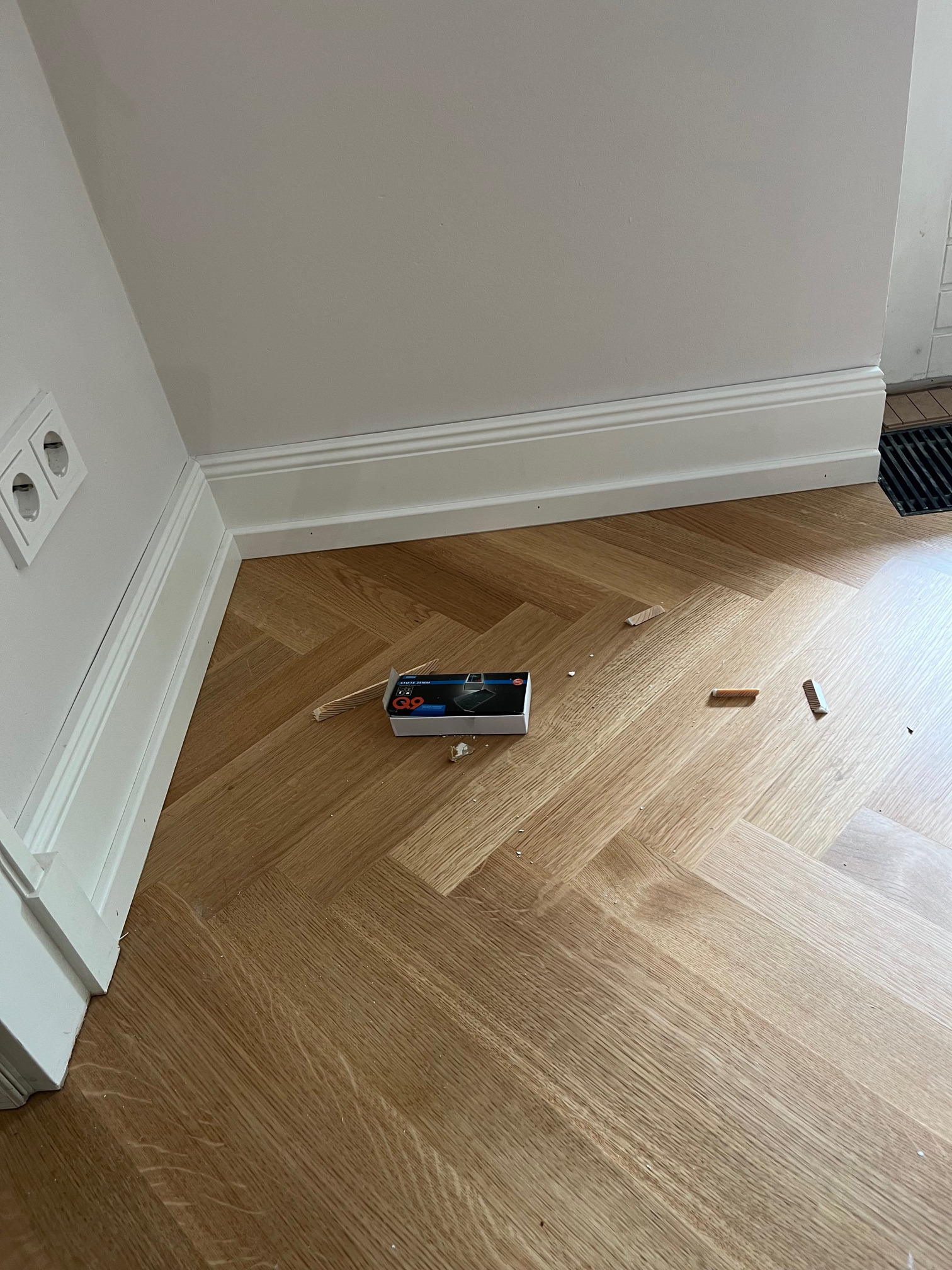 Ułożona podłoga z paneli w jodełkę, przy listwie przypodłogowej. Widoczne narzędzia i resztki paneli. Jasne panele, białe ściany i listwy.