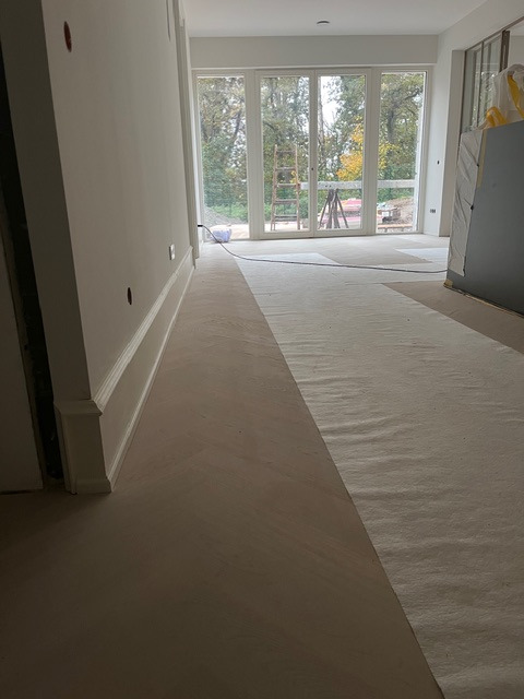 Wnętrze w trakcie wykończenia: świeżo wylana posadzka, zabezpieczona część podłogi, duże okna z widokiem na zewnątrz, drabina w tle. Minimalistyczny wystrój.