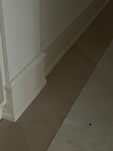 Jasne panele podłogowe ułożone przy białej listwie przypodłogowej z dekoracyjnym frezem. Widok z niskiej perspektywy, podkreślający detale wykończenia.