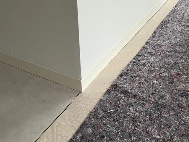 Ochrona podłogi z paneli przed uszkodzeniem podczas remontu, zabezpieczona szarą włókniną. Widoczne białe listwy przypodłogowe i fragment posadzki.