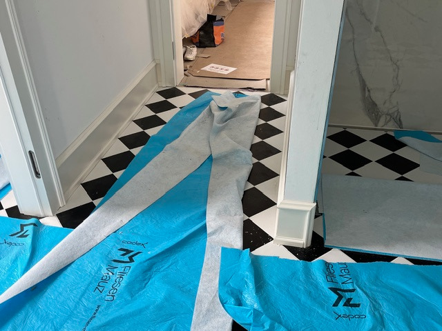 Czarno-biała podłoga w romby, zabezpieczona folią ochronną podczas prac wykończeniowych. Widoczne fragmenty listew przypodłogowych i przejście do innego pomieszczenia.