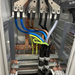 ProfiMajster usługi elektryczne - Wnętrze szafy elektrycznej z profesjonalnie ułożonymi grubymi kablami zasilającymi (niebieski, brązowy, czarny, szary) podłączonymi do miedzianych szyn zbiorczych L1, L2, L3, N, z widocznym...