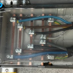 ProfiMajster usługi elektryczne - Wewnętrzne połączenia elektryczne w skrzynce rozdzielczej: miedziane szyny, kolorowe przewody i zaciski, widoczne przez zaparowaną, przezroczystą osłonę.