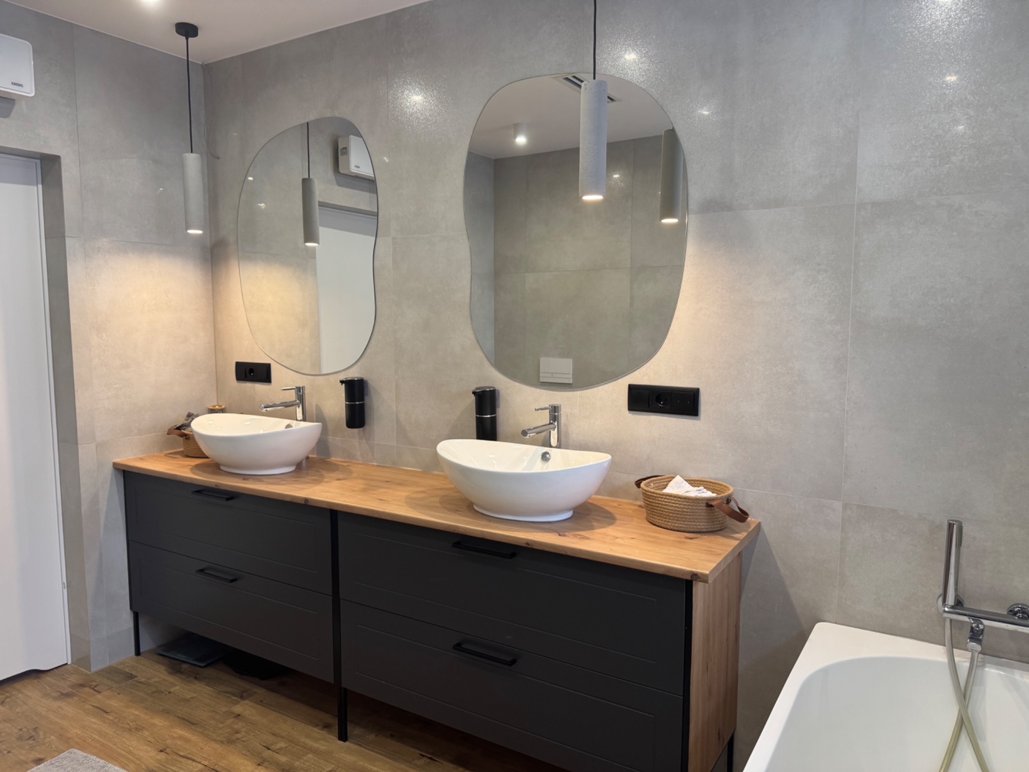 Nowoczesna łazienka z dwoma umywalkami nablatowymi o organicznych kształtach, podwieszanymi lampami i szarą płytką. Uwagę zwraca drewniany blat i czarne szafki.